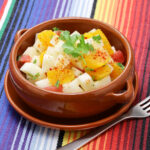 Terra cotta colored bowl filled with a salad composed of oranges and jicama placed on a colorful striped background.