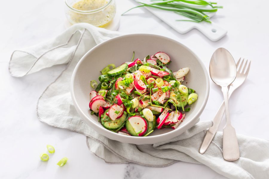 White bowl on a white background filled with a salad composed of radish, cucumber and green onions.