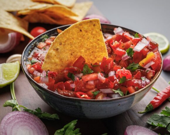 Bowl of tomato salsa topped with tortilla chip set on a wood cutting board with fresh lime, cilantro and chilis.