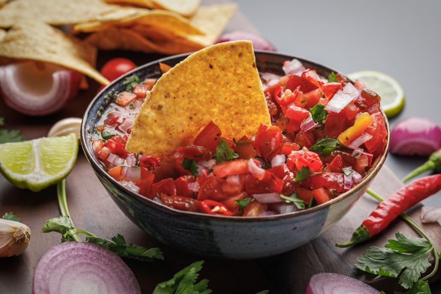 Bowl of tomato salsa topped with tortilla chip set on a wood cutting board with fresh lime, cilantro and chilis.
